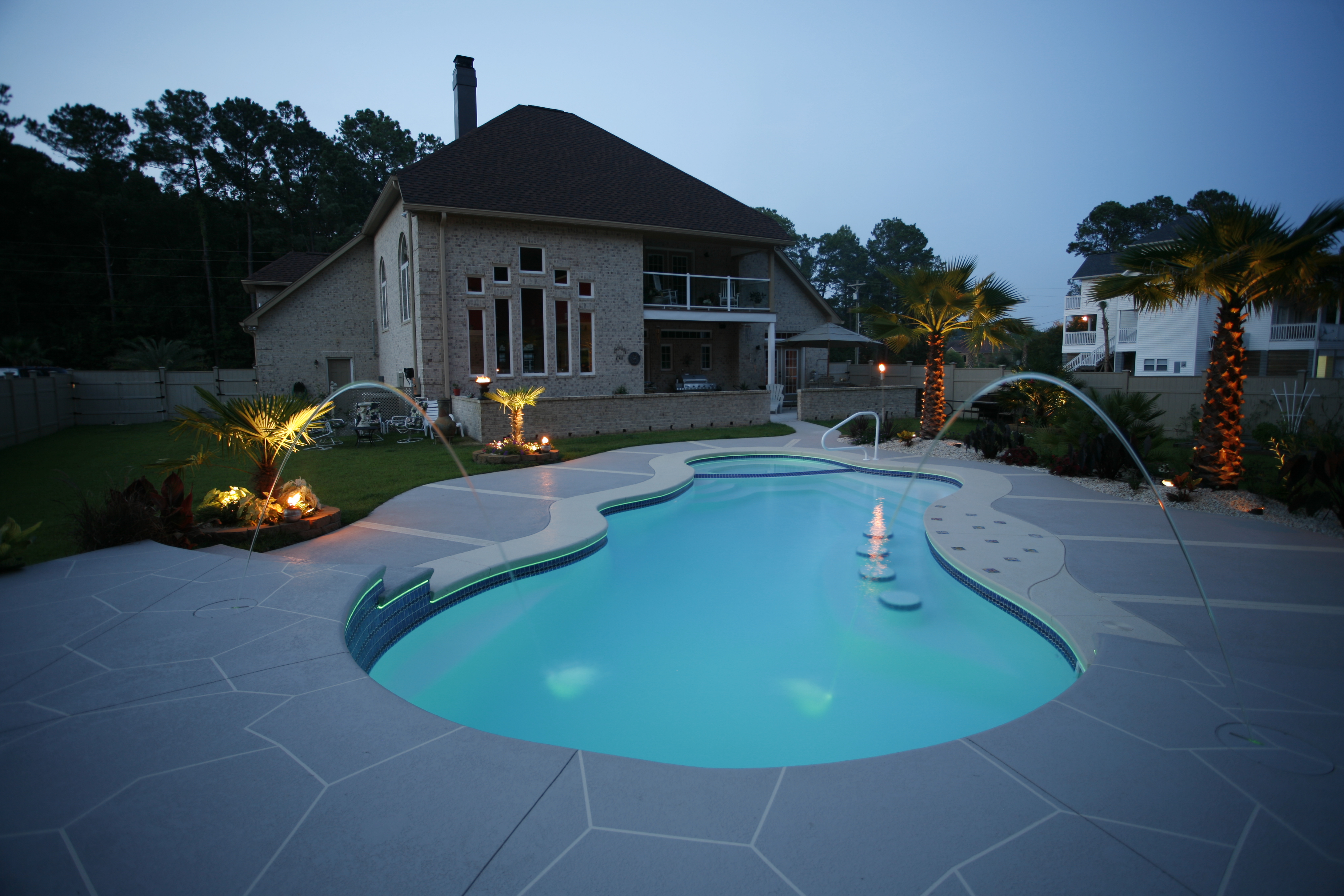 Experience the ultimate in backyard luxury with our fiberglass rectangular pool featuring a tranquil waterfall and stone deck, set amidst lush palm trees and stylish outdoor furniture.
