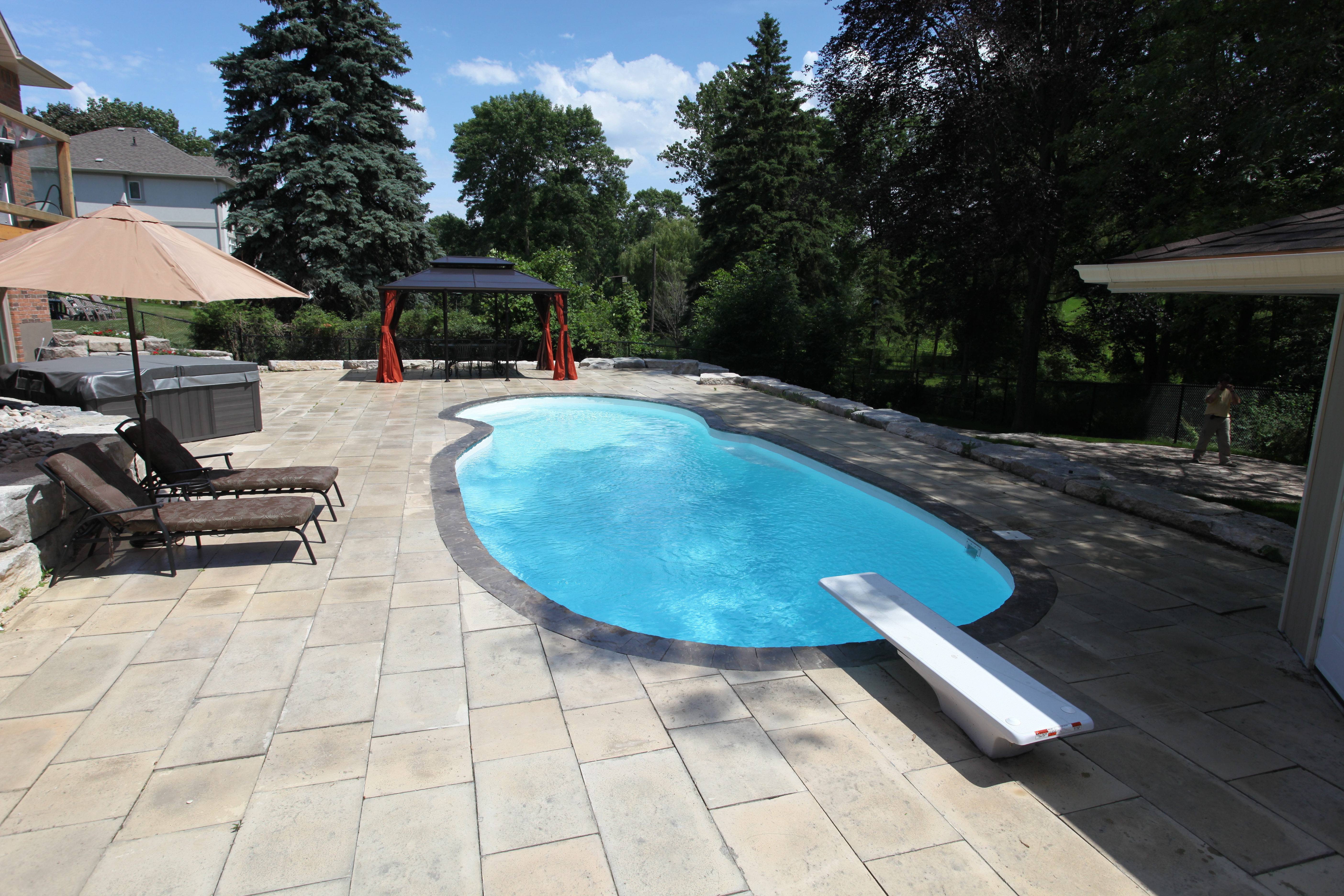Enjoy the ultimate in backyard luxury with this sleek, rectangular fiberglass pool complete with a diving board, surrounded by light-colored pavers and a cozy gazebo—perfect for family gatherings and summer leisure.