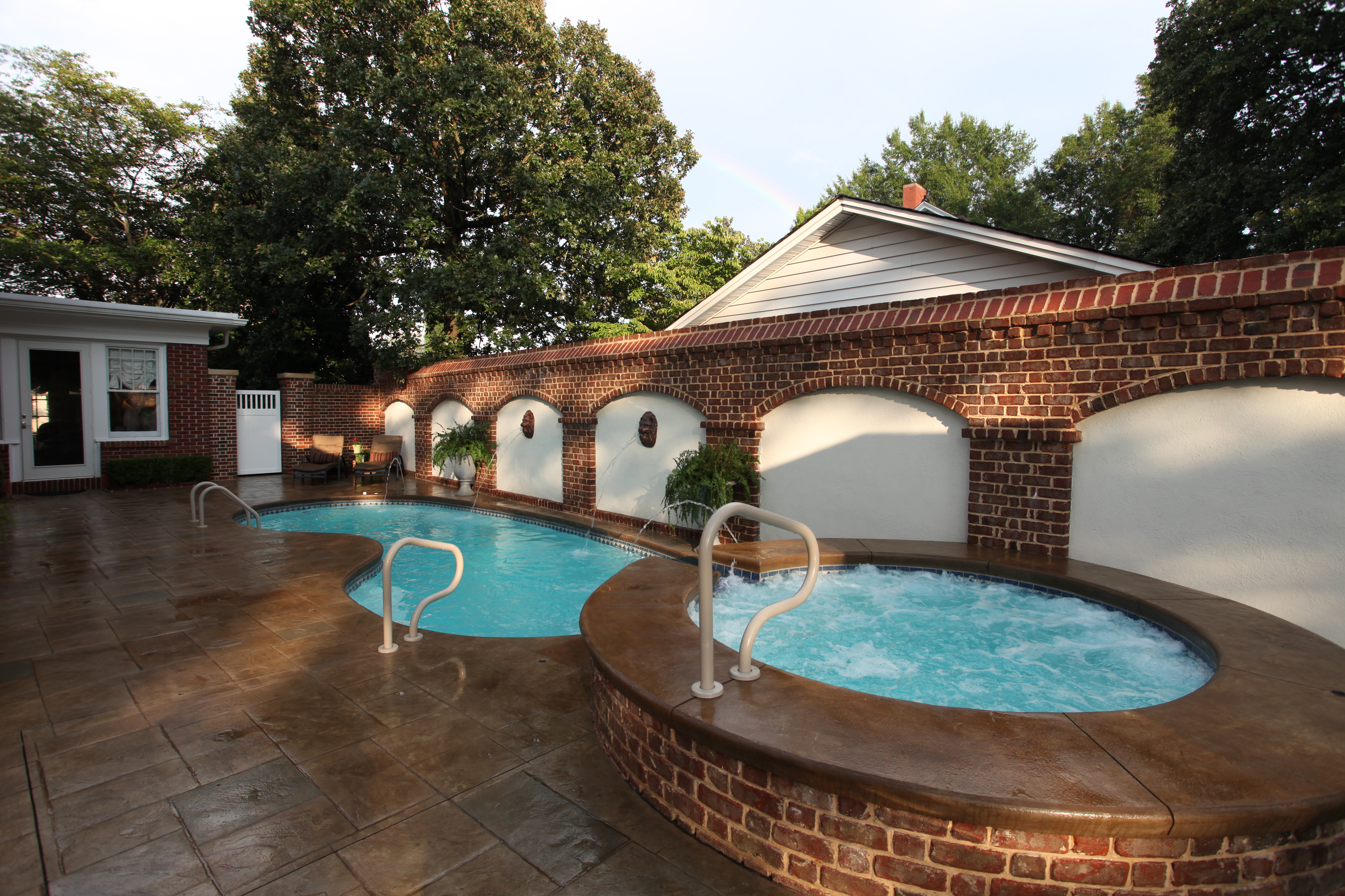 Enjoy a serene summer retreat with our fiberglass pool and hot tub, surrounded by lush greenery and a classic brick wall.