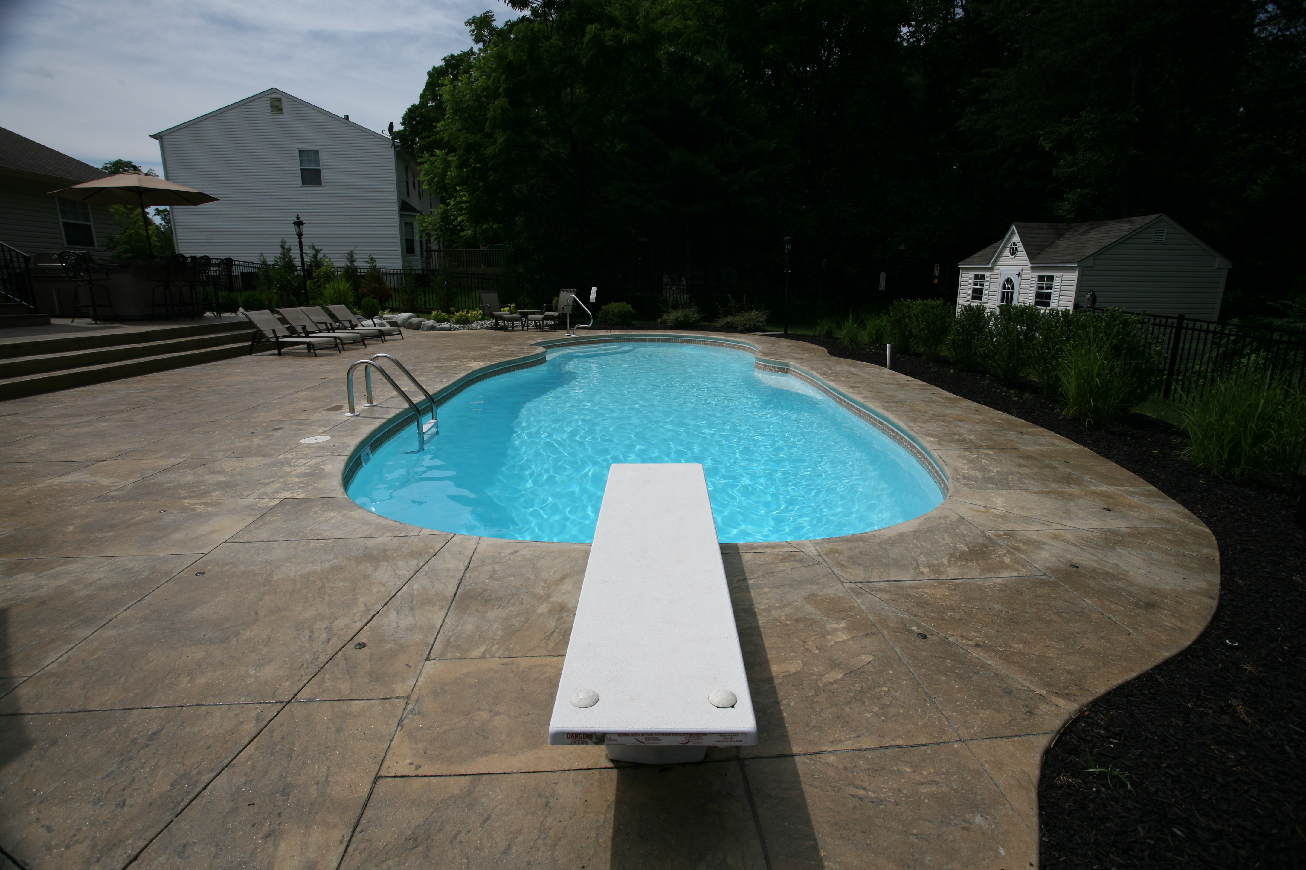 Enjoy a sparkling fiberglass pool in a sunny backyard, complete with a white diving board and cozy seating—perfect for summer relaxation.