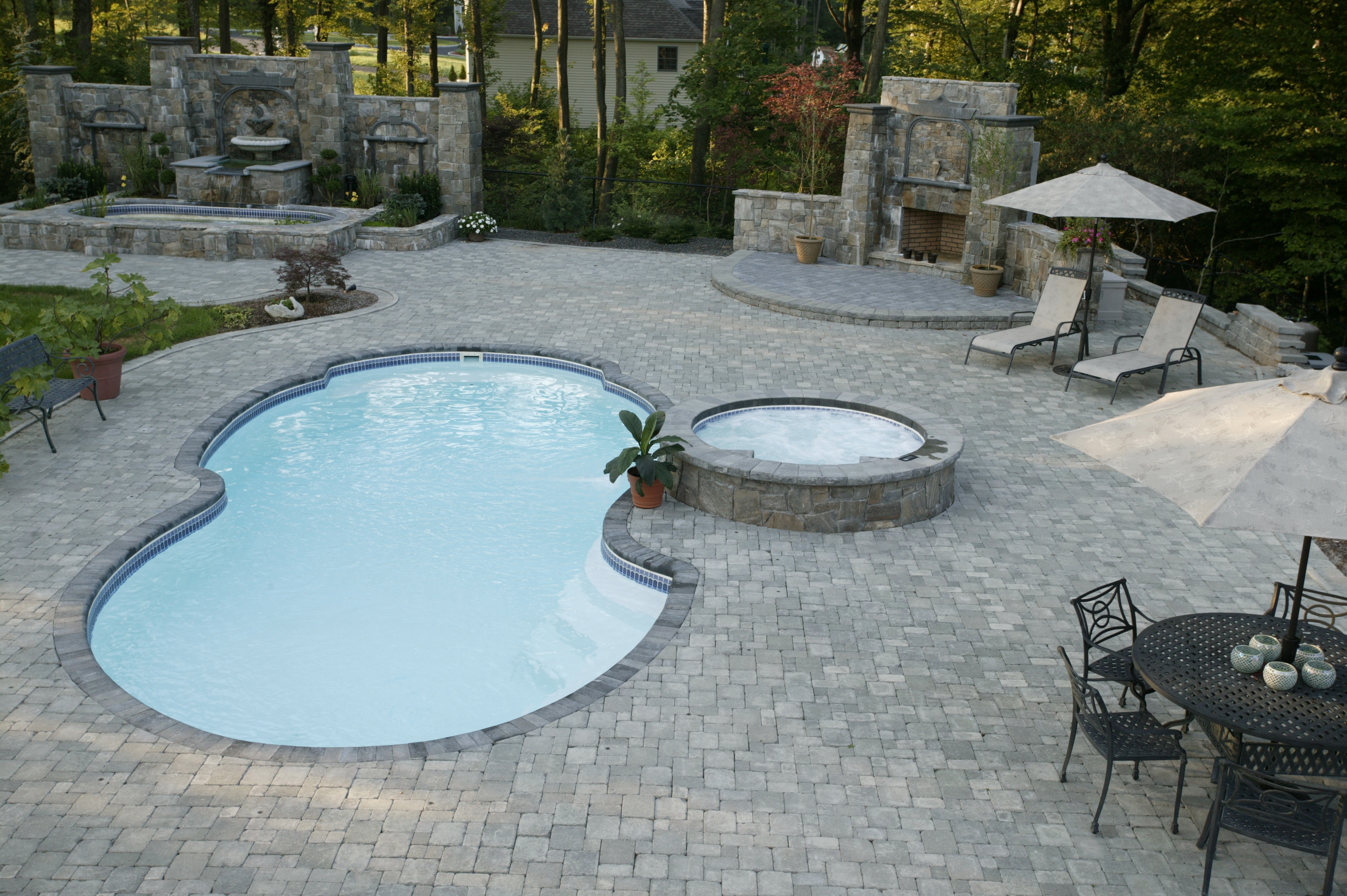 Enjoy a luxurious, sunlit backyard oasis featuring a spacious fiberglass in-ground pool surrounded by a stone patio, hot tubs, and lush greenery.