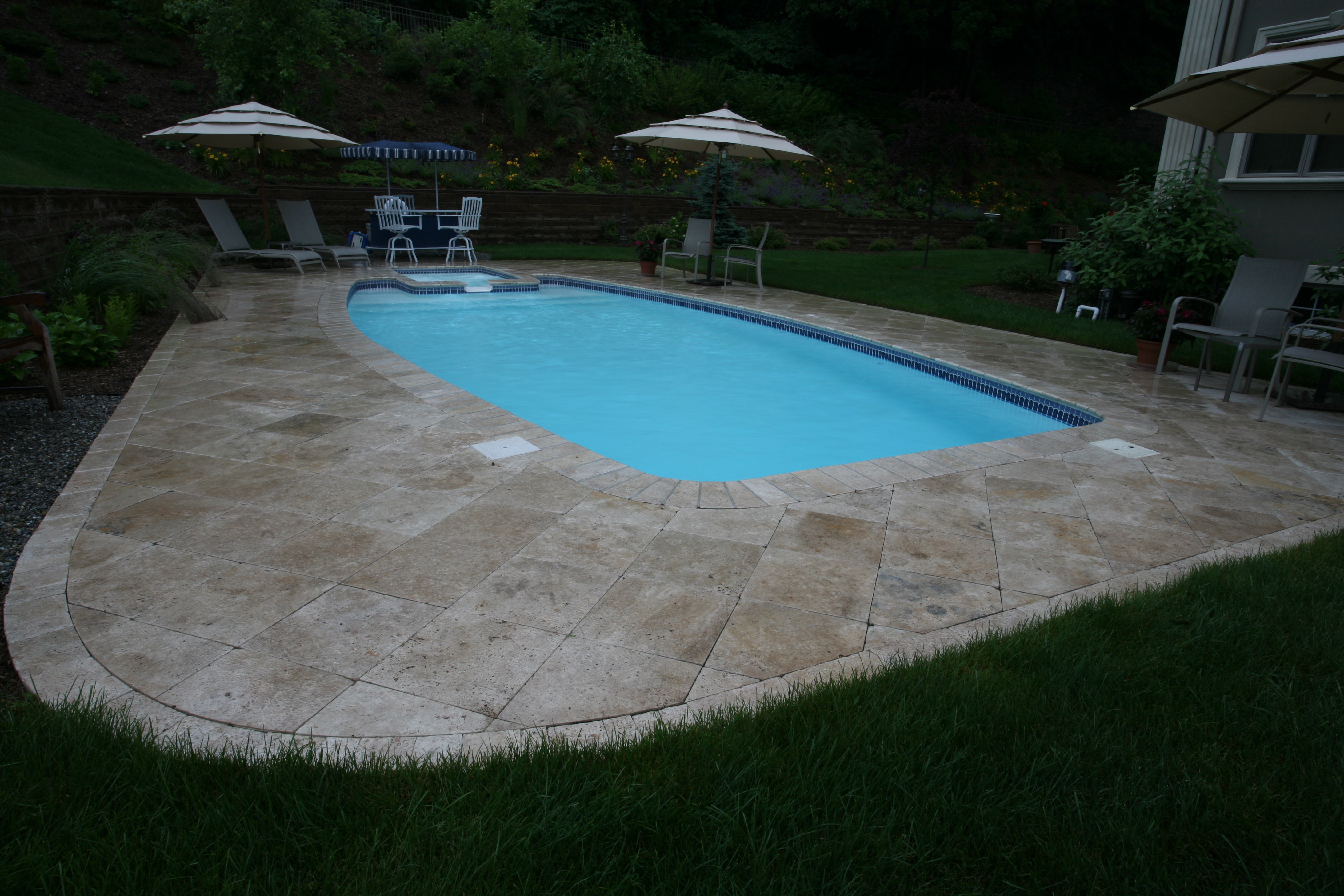 Experience tranquil evenings in your own backyard with San Juan’s fiberglass kidney-shaped pool, set amid light stone pavers and lush greenery.
