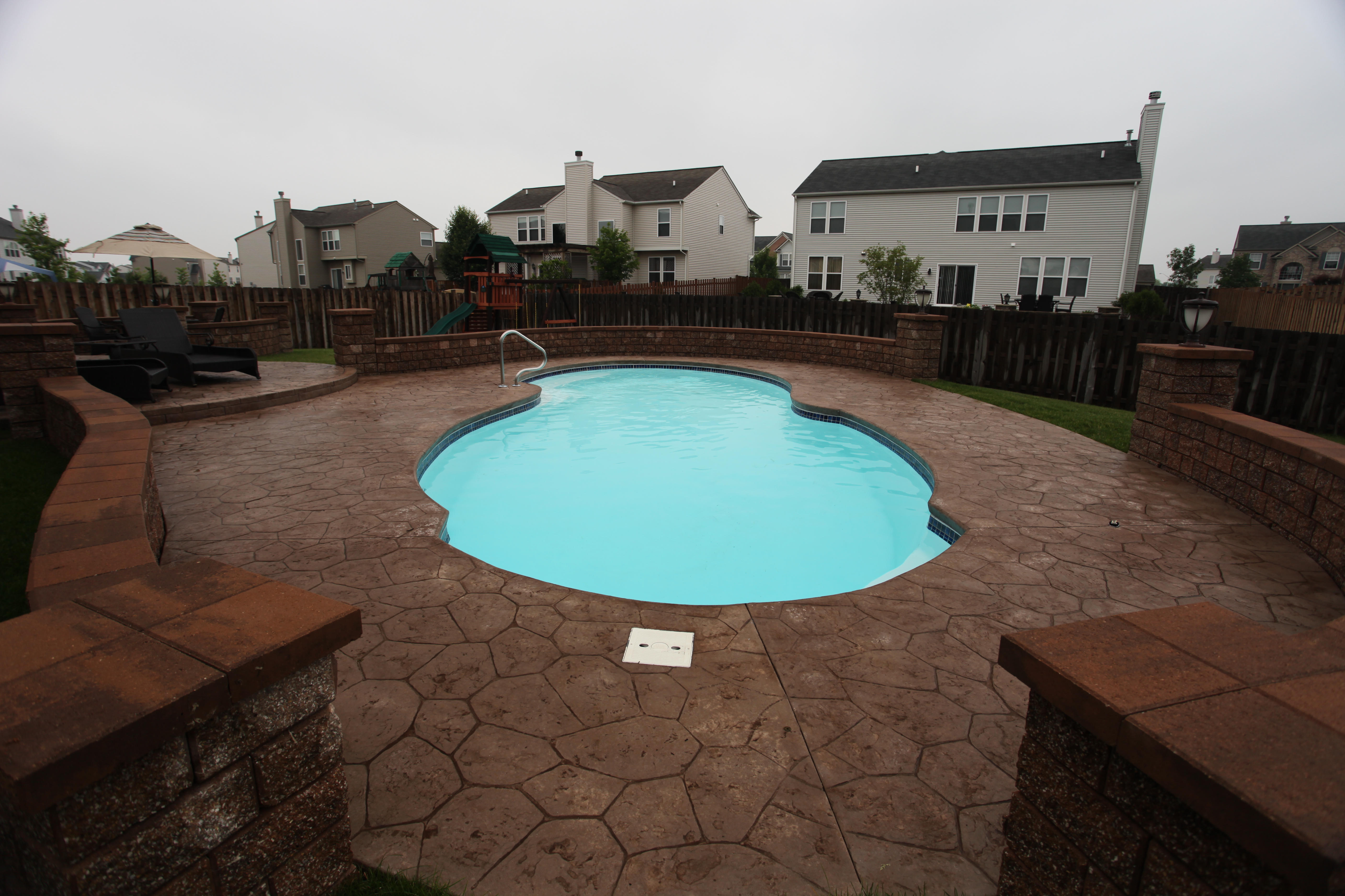 A serene backyard showcases a sleek fiberglass kidney-shaped pool set amid a warm stamped-concrete deck and subtle overcast lighting.