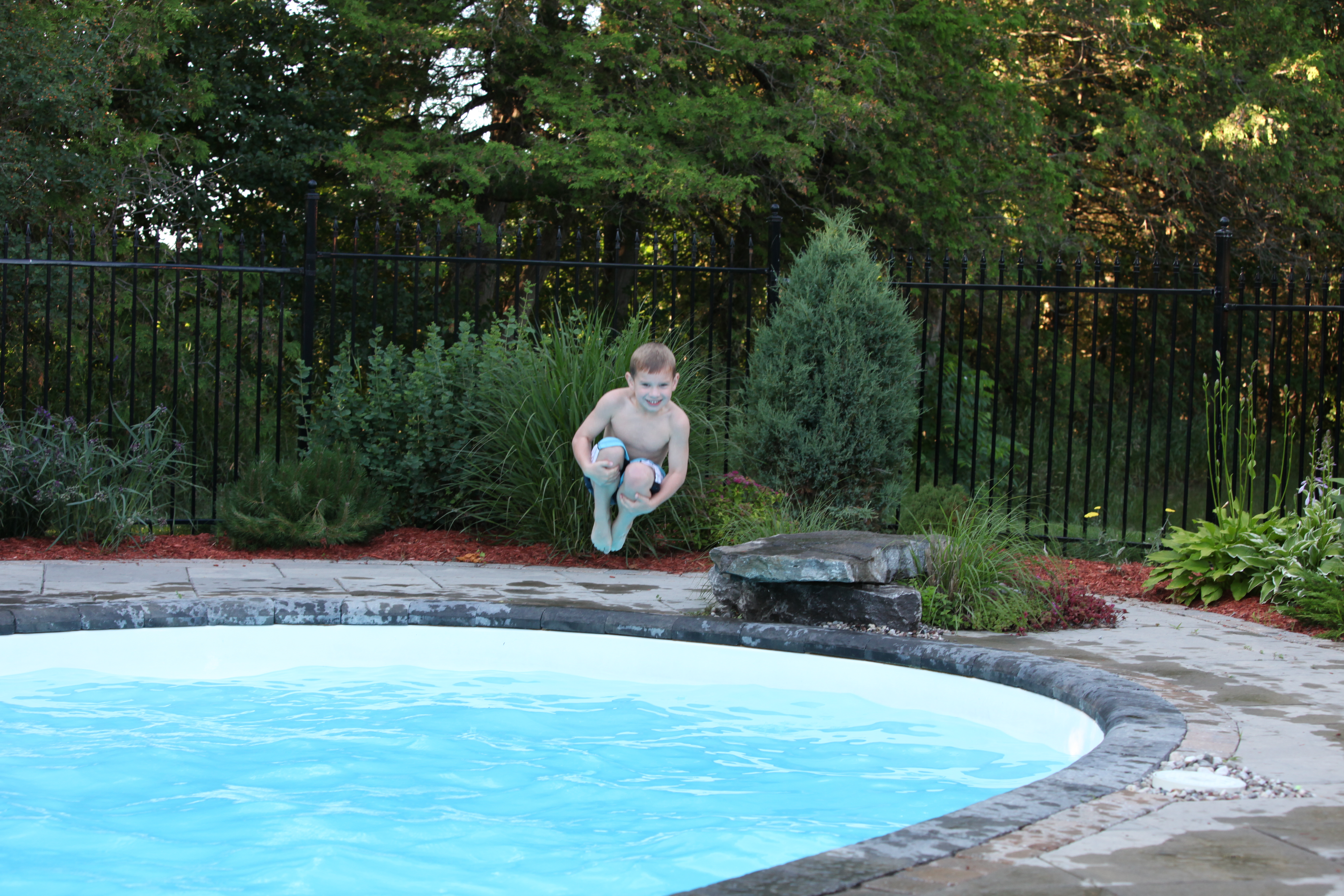 A joyful boy leaps over a sparkling fiberglass pool set in a sun-kissed backyard, showcasing crystal-clear water and vibrant landscaping.