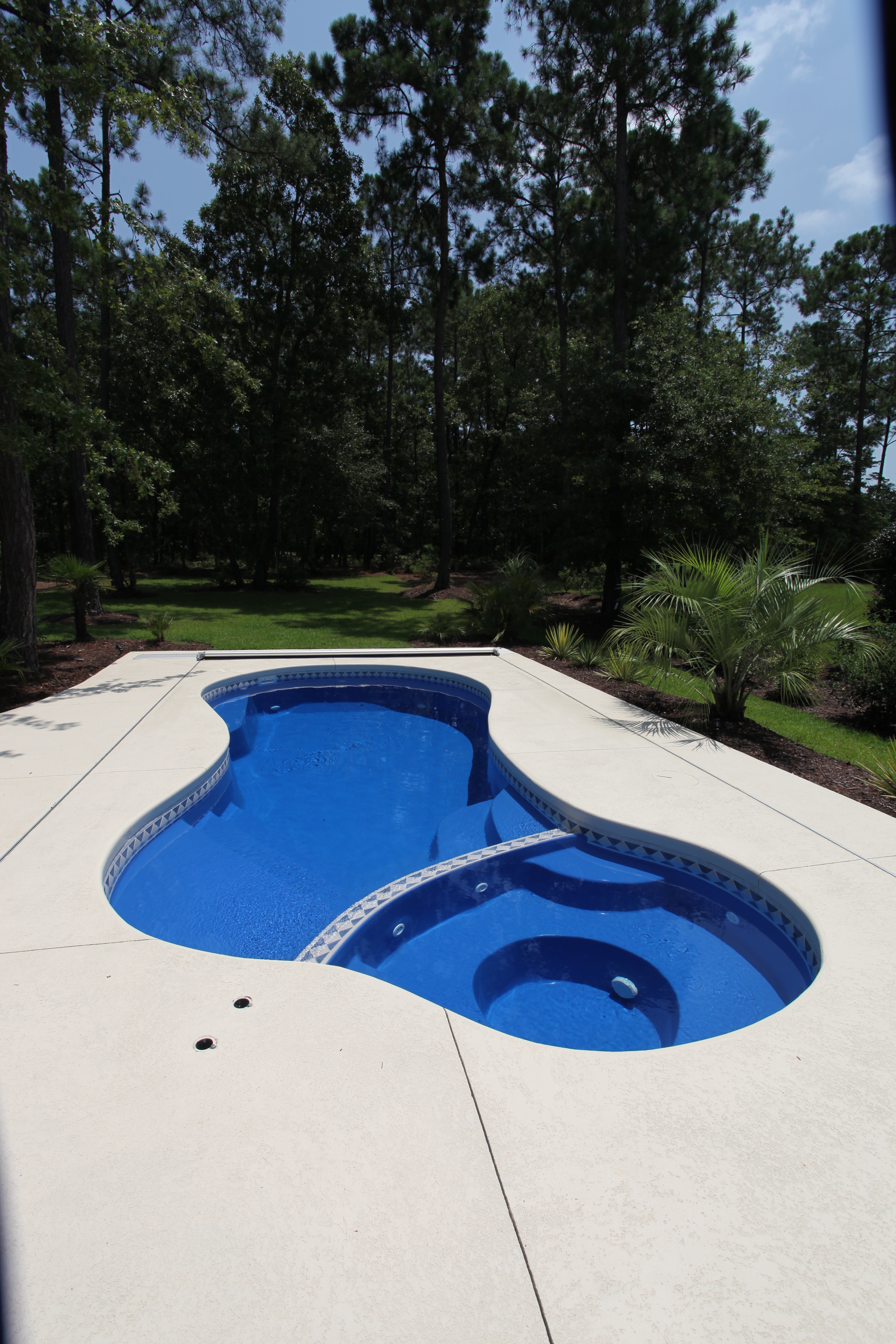 Experience tranquil luxury with this sleek fiberglass pool, featuring a curvilinear design and an integrated spa set amidst a serene wooded backyard.