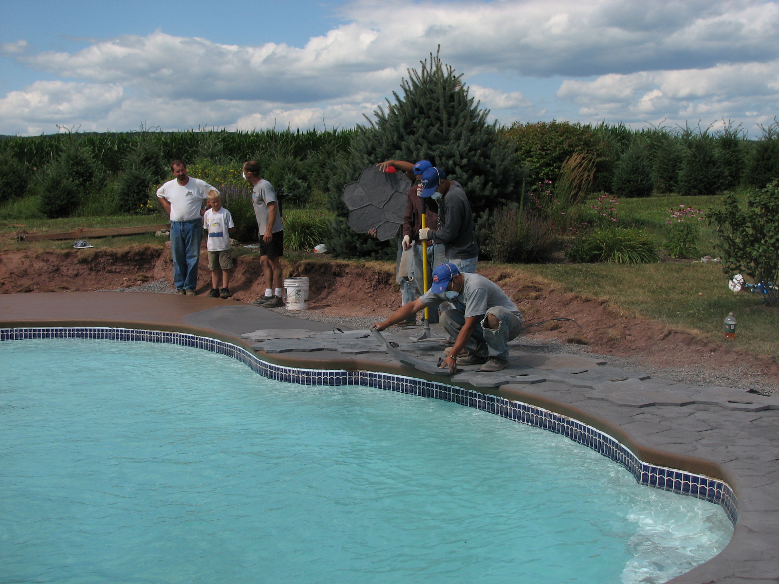 A newly installed fiberglass pool sparkles under a bright sky as workers finish the surrounding deck, showcasing San Juan’s quality and natural backyard integration.
