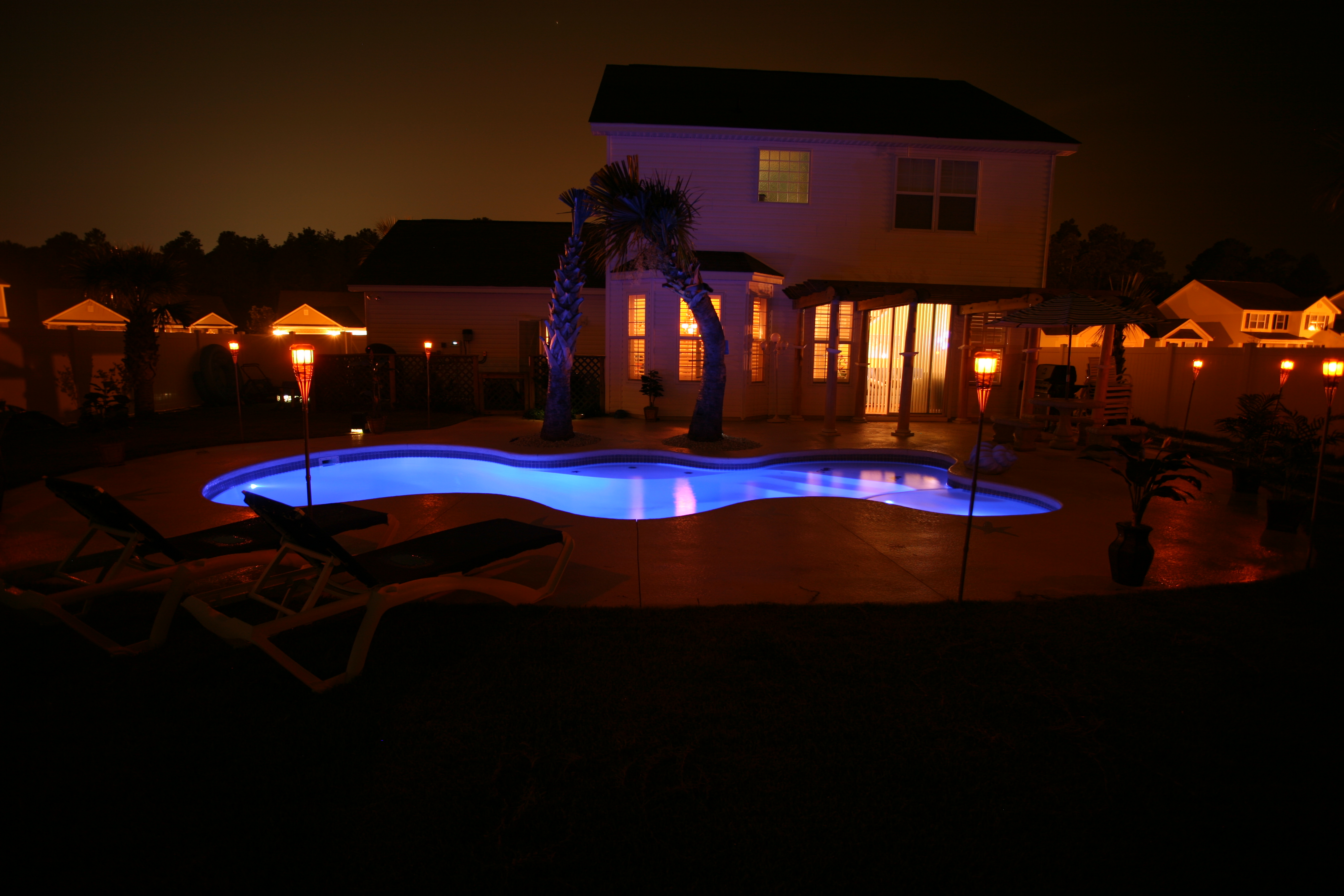 A serene desert oasis at night, featuring a modern inground pool as the centerpiece of a relaxing backyard retreat. The warm glow of the pool lights creates a welcoming ambiance, perfect for a summer evening.