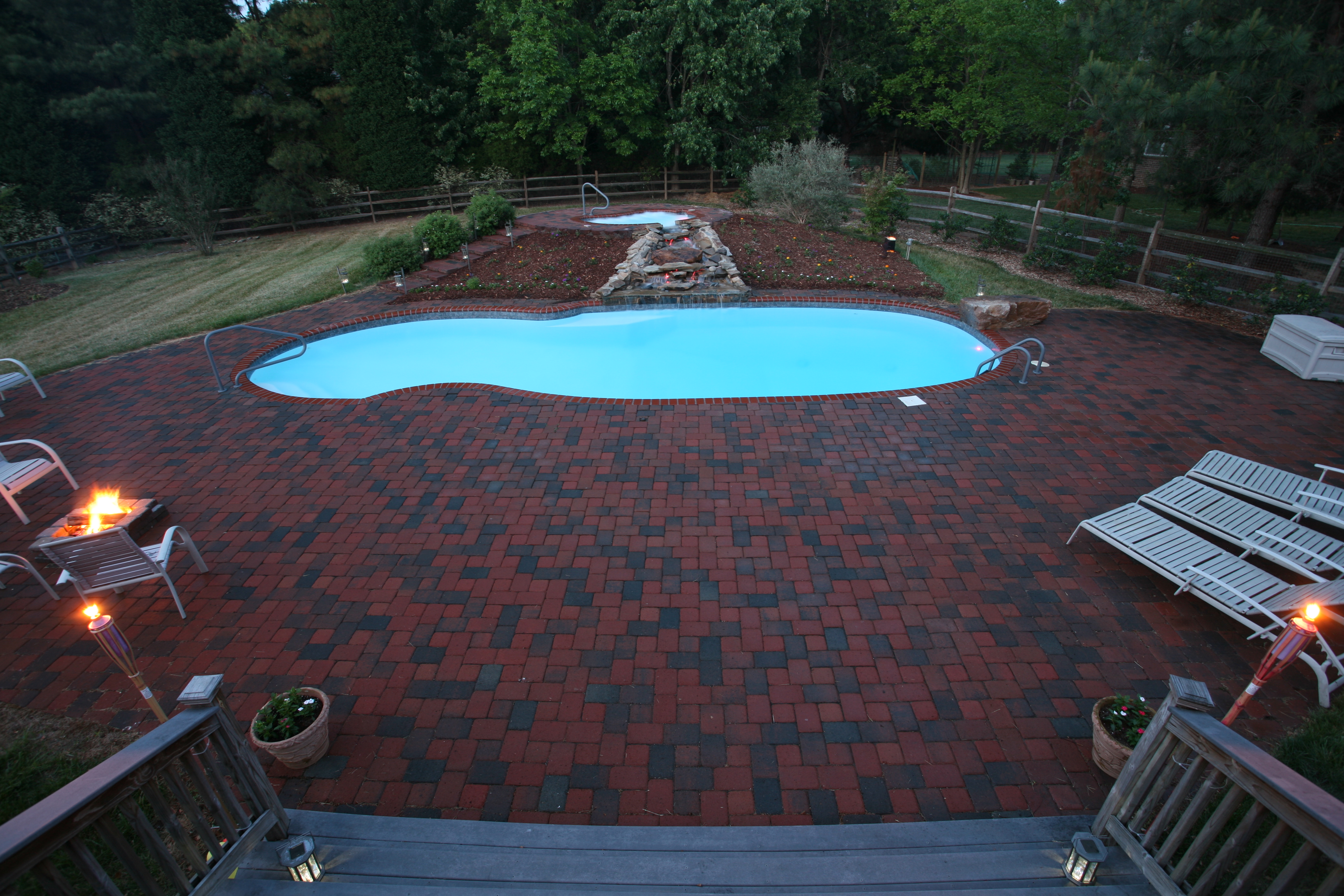 Experience tranquility with this inviting fiberglass pool scene, featuring a vibrant blue pool surrounded by a red brick patio and lush greenery.
