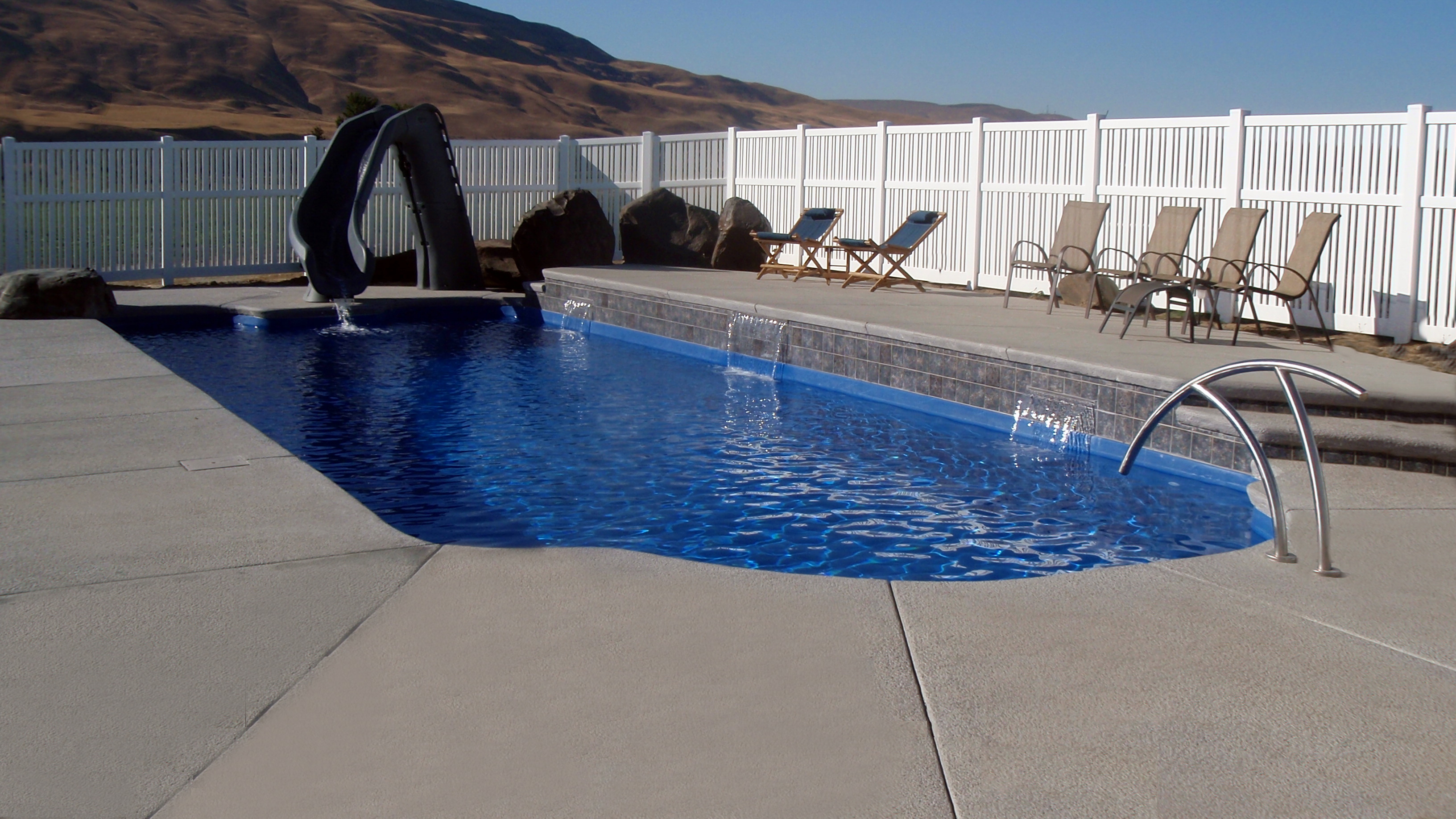 Experience tranquility with this vibrant blue fiberglass plunge pool, surrounded by a clean concrete deck and lounge chairs in a serene backdrop.