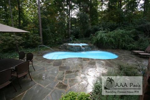 Fiberglass pool in a forested backyard with stone patio and hot tub.